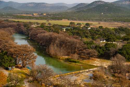 A scenic view of a river winding through trees and hills, with a clear sky and distant mountains in the background.