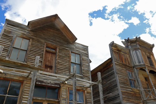Two weathered wooden buildings with a blue sky and clouds in the background, showcasing a historic ghost town.