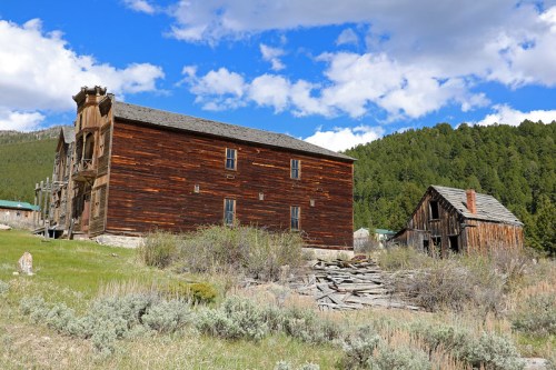 Two weathered wooden buildings in a grassy area, surrounded by trees and under a blue sky with clouds.