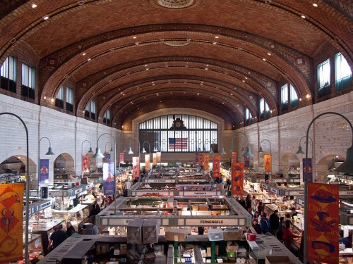 A bustling market interior with high arched ceilings, rows of vendor stalls, and colorful banners hanging from the walls.