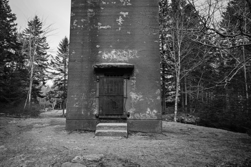 A weathered building with a barred door, surrounded by trees in a black and white landscape.