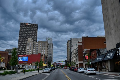 A city street lined with buildings and parked cars under a cloudy sky, showcasing urban architecture and a vibrant atmosphere.