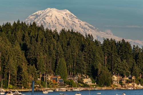 A scenic view of a mountain with snow-capped peaks, surrounded by lush forests and a calm lake dotted with boats.