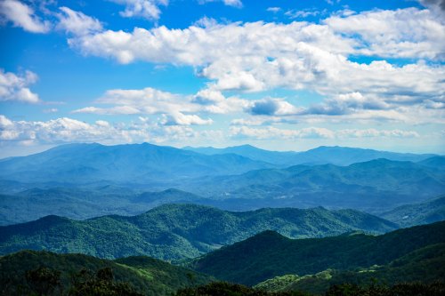 A panoramic view of rolling green mountains under a bright blue sky with fluffy white clouds.