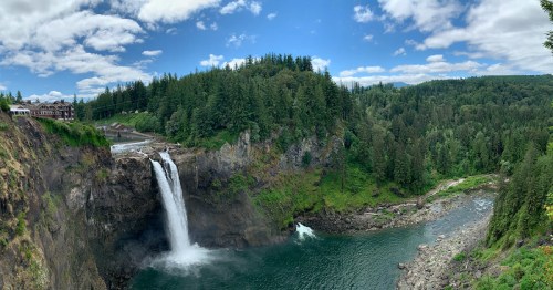 A panoramic view of a waterfall cascading into a river, surrounded by lush green trees and a blue sky with clouds.