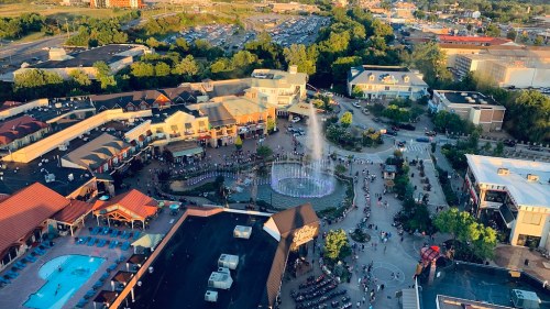 Aerial view of a bustling shopping area with a fountain, surrounded by trees and parked cars in the background.