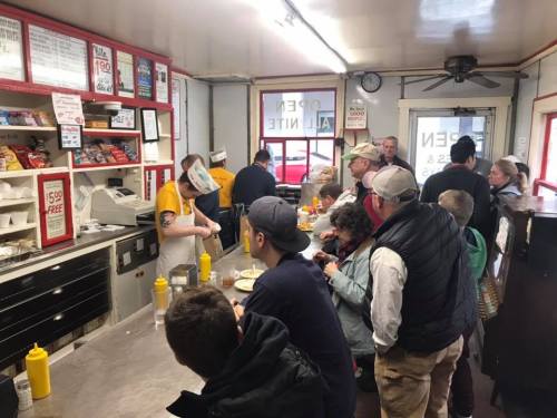 A busy diner with customers waiting in line, a staff member preparing food behind the counter, and condiments on the table.