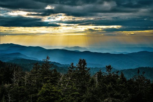 A scenic view of layered mountains under a dramatic sky, with sunlight breaking through clouds and illuminating the landscape.