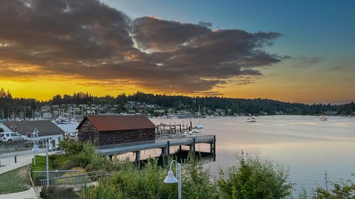 A serene waterfront scene at sunset, featuring a dock, boats, and a rustic building against a colorful sky.