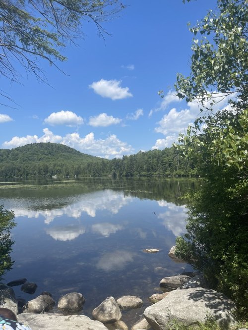 A serene lake reflects a blue sky and clouds, surrounded by green trees and a distant hill. Rocks line the shore.