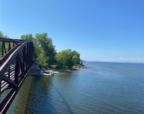 A metal bridge arches over a calm lake, surrounded by green trees and a clear blue sky.