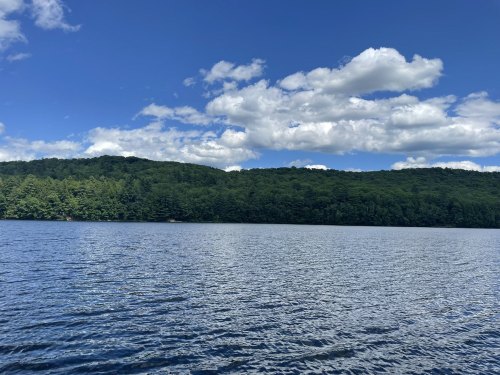A serene lake surrounded by lush green hills under a bright blue sky with fluffy white clouds.