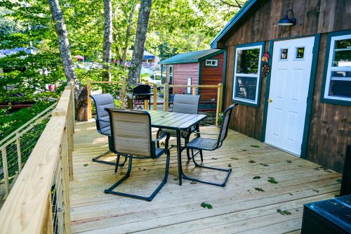 A wooden deck with a table and four chairs, surrounded by trees and small cabins in the background.