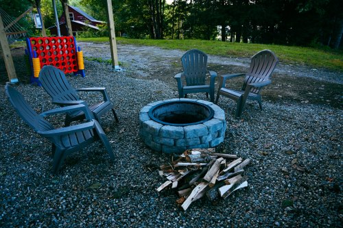 A circular stone fire pit surrounded by four gray chairs and a pile of firewood on a gravel surface.