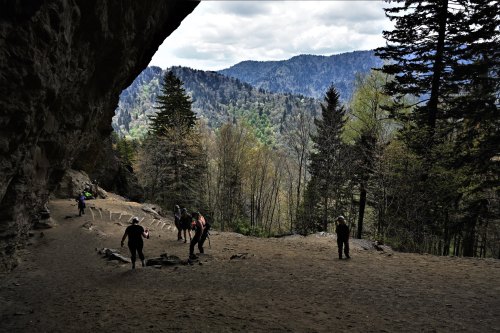 A group of people exploring a cave entrance with a scenic mountain view and lush trees in the background.
