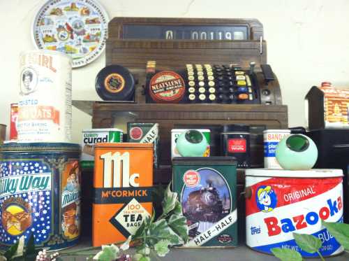 A vintage display featuring an old cash register, various retro food cans, and decorative items on a shelf.