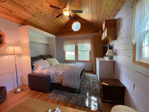 Cozy bedroom with wooden walls, a queen bed, windows, a ceiling fan, and a small dresser. Natural light fills the space.