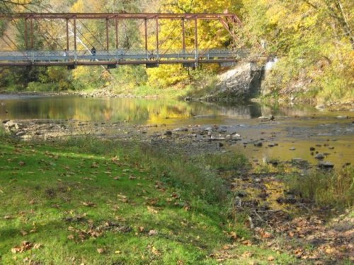 A rustic bridge spans a calm river, surrounded by autumn foliage and a grassy bank with scattered leaves.
