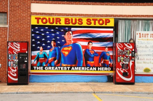 Mural of superheroes in front of a brick wall, featuring a large American flag and Coca-Cola vending machines.