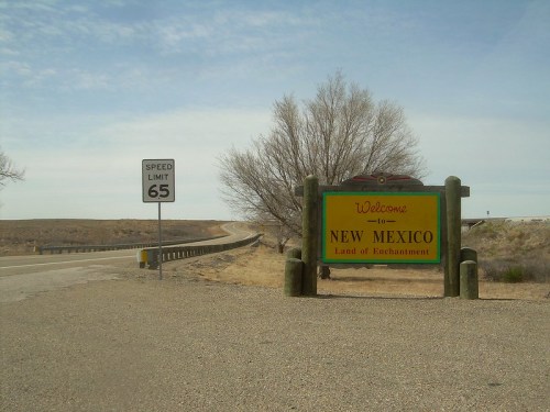 Welcome sign for New Mexico, "Land of Enchantment," with a speed limit sign and a barren landscape in the background.