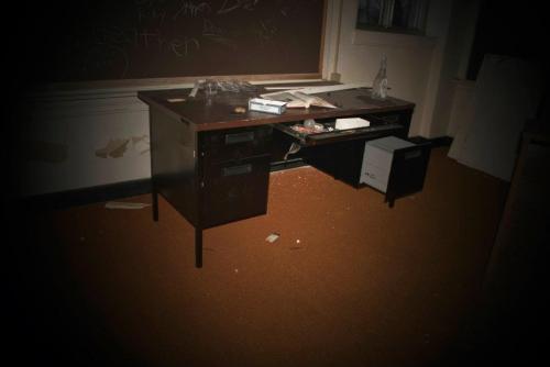 An abandoned classroom with a messy desk, scattered papers, and debris on the floor, dimly lit.