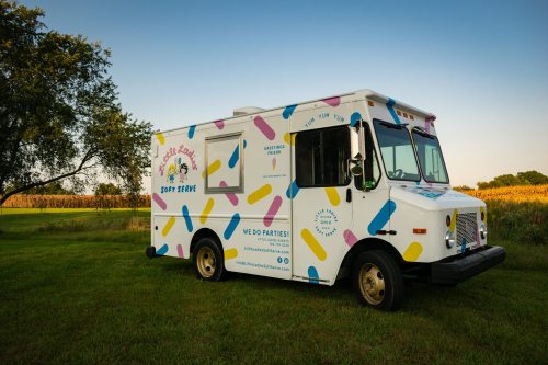 A colorful ice cream truck parked in a grassy field, surrounded by trees and a cornfield under a clear blue sky.