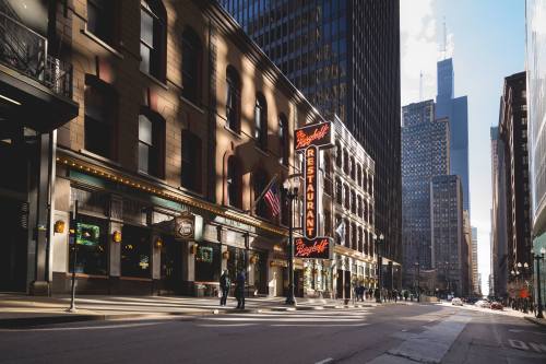 A city street with a restaurant sign, tall buildings, and pedestrians under a clear sky.