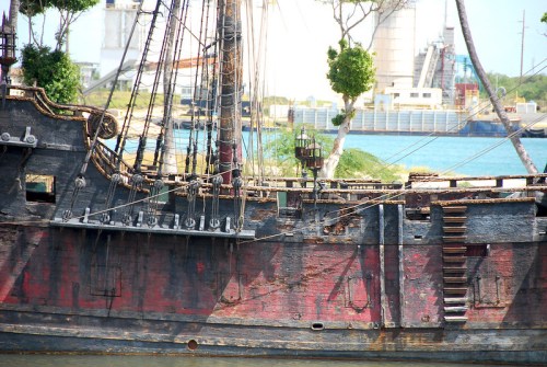 A weathered pirate ship with peeling paint, moored near a harbor with industrial buildings in the background.