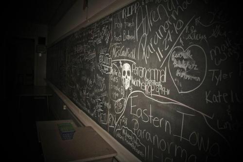 A dimly lit classroom chalkboard filled with various writings, drawings, and a prominent skull illustration.