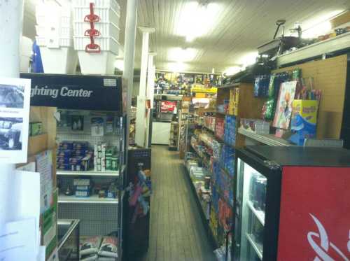 A narrow aisle in a small store lined with shelves of various products, including snacks and household items.