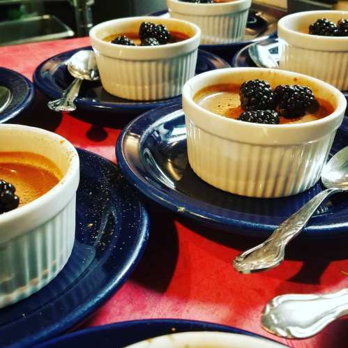 Several ramekins of dessert topped with blackberries, arranged on blue plates with silver spoons beside them.