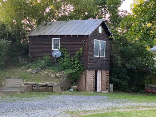 A small, rustic wooden house with a metal roof, surrounded by greenery and gravel, featuring a satellite dish.