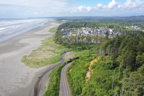 Aerial view of a coastal town with sandy beach, lush greenery, and winding road along the shoreline.