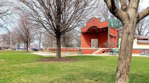 A red outdoor stage in a park, surrounded by bare trees and green grass, with a playground visible in the background.