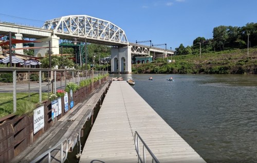 A wooden dock extends over a calm river, with a bridge in the background and people kayaking nearby under a clear blue sky.
