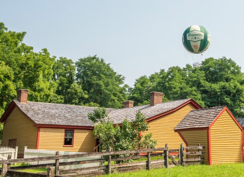 A yellow house with red trim and a wooden fence, with a green and white hot air balloon floating above it.
