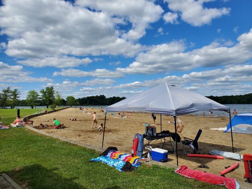 A sandy beach with people sunbathing and playing, under a blue sky with fluffy clouds and a shaded tent.