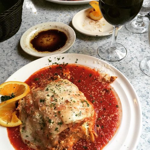 A plate of lasagna topped with cheese and marinara sauce, garnished with parsley and orange slices, alongside a glass of red wine.