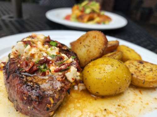 A plate featuring a grilled steak topped with sun-dried tomatoes and herbs, alongside roasted potatoes and yellow squash.