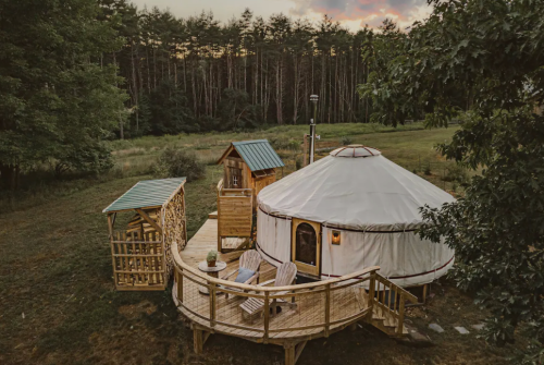 A cozy yurt with a wooden deck, surrounded by trees and a grassy field, at sunset.