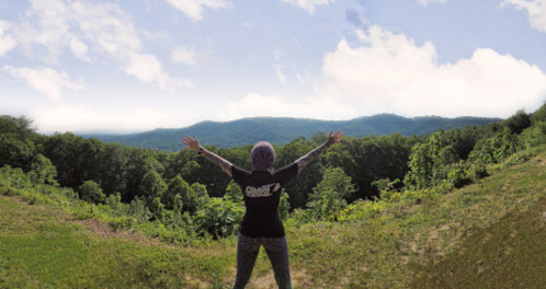 Person with arms outstretched, standing on a hillside overlooking lush green mountains and a blue sky.