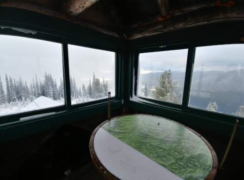 A cozy lookout tower with large windows offering a view of snowy mountains and a green map on the table.