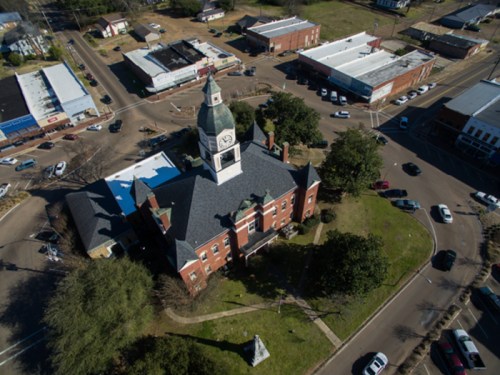 Aerial view of a historic building with a clock tower, surrounded by trees and small shops in a town setting.