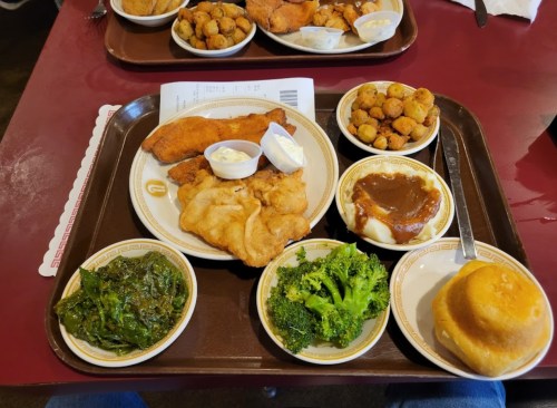 A tray with fried fish, sides of greens, broccoli, okra, and a roll, accompanied by dipping sauces.