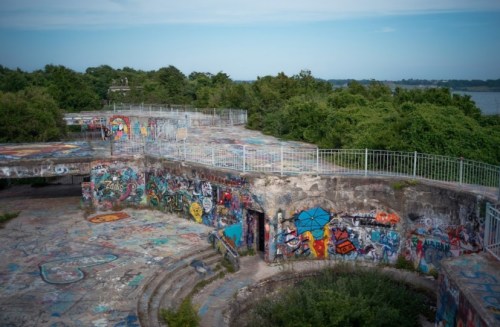 Aerial view of a graffiti-covered, abandoned structure surrounded by lush greenery and a body of water in the distance.