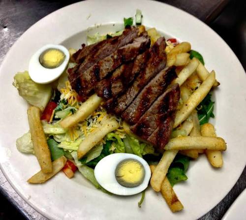 A plate of salad topped with grilled steak, French fries, and sliced hard-boiled eggs.