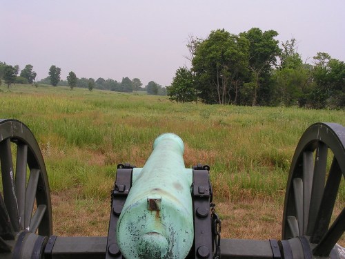 View from behind a historic cannon, overlooking a grassy field and trees in the distance under a hazy sky.