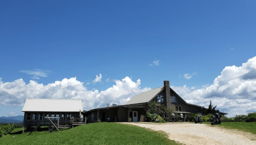 A rustic building with a stone chimney under a bright blue sky, surrounded by green grass and distant mountains.