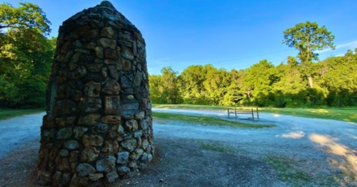 A stone monument stands beside a gravel path, surrounded by lush greenery and trees under a clear blue sky.