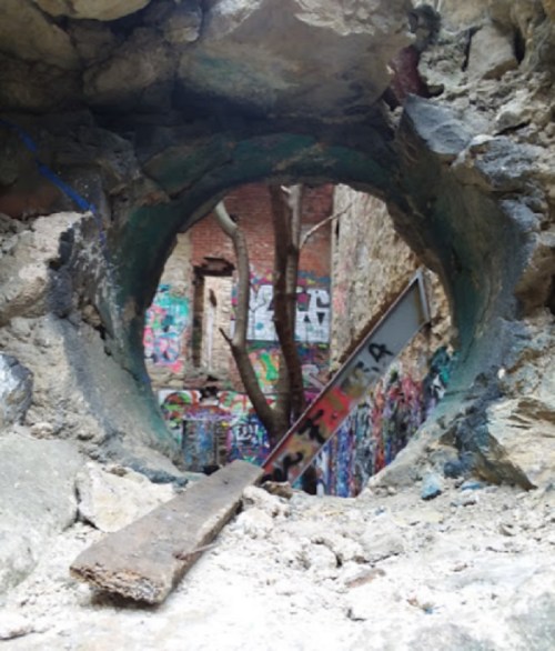 A circular opening in a stone wall reveals a colorful, graffiti-covered space with a tree growing inside.